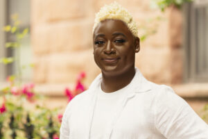 Picture of Lady Anderson. Lady is a Black woman with short curly bleached blonde hair. She is wearing a white button up over a white t shirt and posing in front of some pink flowers. She has a soft smile on her face.