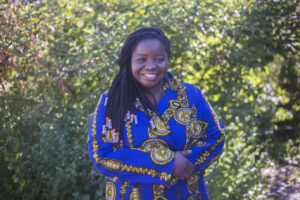 Picture of Valeriana. An Angolan woman with long sideswept braids wearing a cobalt blue print dress poses in front of a lush outdoor backdrop. She has a warm and kind smile.