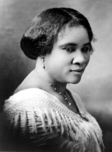 Black and white photo of Madam CJ Walker. Antique photo portrait of a Black woman with her hair gathered at her neck wearing a graceful white gown and jewelry and looking down with a gentle expression