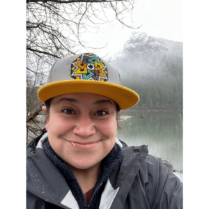 Selfie of Mija an Indigenous person wearing a baseball cap and a raincoat. They are standing in front of a mountainside lake. They look directly into the camera with a big grin and an amused look on their face.