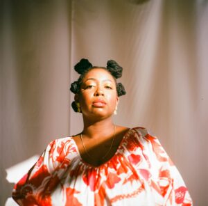 Picture of Joy Alise Davis a Black woman seated in a ray of golden sun. She wears her hair in knots with gold jewelry and a red and white print top. She looks downwards at the camera with an unbothered expression.