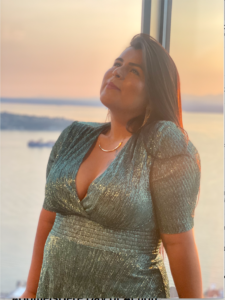 Picture of Monserrat a brown woman with long dark hair wearing a sparkling aqua dress. She is leaning back against a window in golden hour light looking up and away from the camera. She has a dreamy look on her face.