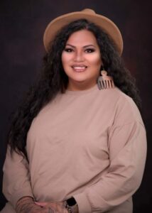 Picture of Taffy a Samoan woman with long black hair. She wears a taupe top wood earrings and a beige hat. She is smiling calmly.