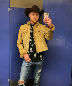Picture of Wesley a white man posing in front of a blue backdrop. He wears a black cowboy hat a yellow jacket a black shirt and jeans with glasses.