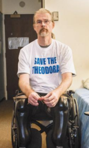 Picture of Shawn Walton a white man in a wheelchair with prosthetic legs. He has a beard and wears glasses and is wearing a white t shirt with blue lettering reading Save the Theodora
