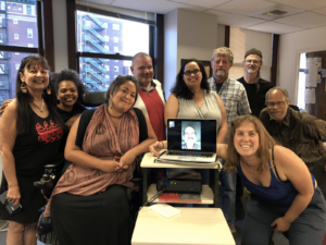 Picture of DTAT members gathered at an office. A group of white Black and brown people gathered together around a computer with a man on zoom displayed. They smile at the camera.