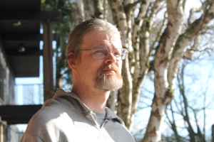 Picture of Shawn Walton a white man with a beard and glasses with a solemn expression. He stands outside on a sunny day in front of a tree
