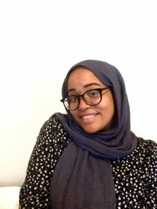 Photo of Aisha. A mixed Black Muslim girl smiling into the camera with a playful expression and one shoulder up in a shrug. She wears a grey headscarf black glasses and a black flower print top.