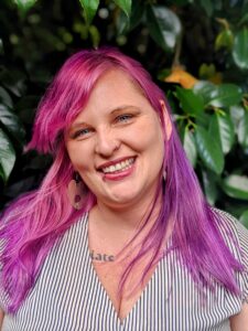 Picture of Amy a white woman with pink and purple hair in front of a green leafy backdrop. She has a carefree smile and is wearing a black and white striped top.