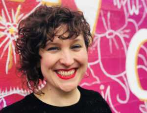 Picture of Mercedes a white woman with short curly brown hair and a wide grin. She is posing in front of a bright pink backdrop and is wearing a black top.