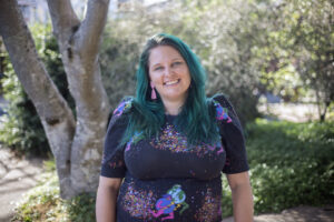 Picture of Amy. A white woman with bright teal hair and a black print top stands in front of a lush outdoor backdrop. She looks directly into the camera with an infectious smile