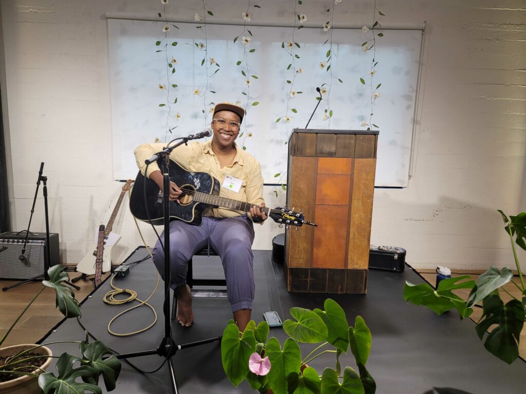 Picture of Elisha a nonbinary Black person playing guitar on a stage. They are surrounded by plants and a backdrop of white paper flowers.