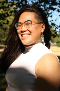 Picture of Florence a Chinse person smiling in the sun. Flo has long black hair with one side shaved and they are wearing colorful earrings a white top and glasses.