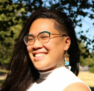 Picture of Florence a Chinse person smiling in the sun. Flo has long black hair with one side shaved and they are wearing colorful earrings a white top and glasses.