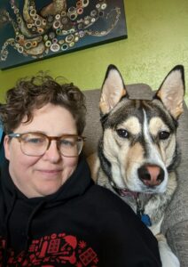 Picture of Bdub a white person with brown hair wearing glasses and a black sweatshirt posing with their husky mix Brioche who is very very cute
