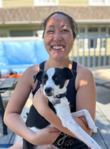 Picture of Emily Chu. An Asian woman wearing a black tank top and holding a medium black and white dog is picture. She is smiling with light and shadows on her face.