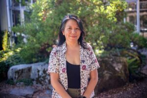Picture of Precious a smiling Filipino woman with black hair. She is posing in a sunlit garden.