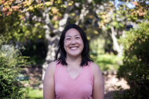 Picture of Emily a smiling East Asian woman wearing a pink top posing in a sunlit garden.