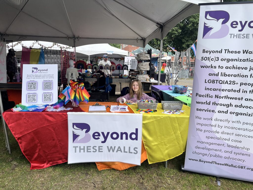A Beyond These Walls community outreach booth at a Pride event displays rainbow flags, educational materials, and colorful table coverings. A child sits behind the table helping welcome visitors.