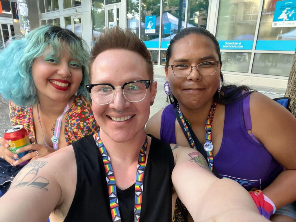 Three smiling people pose for a selfie outdoors at an LGBTQ+ event. They wear rainbow lanyards and colorful clothing while standing on a city sidewalk.