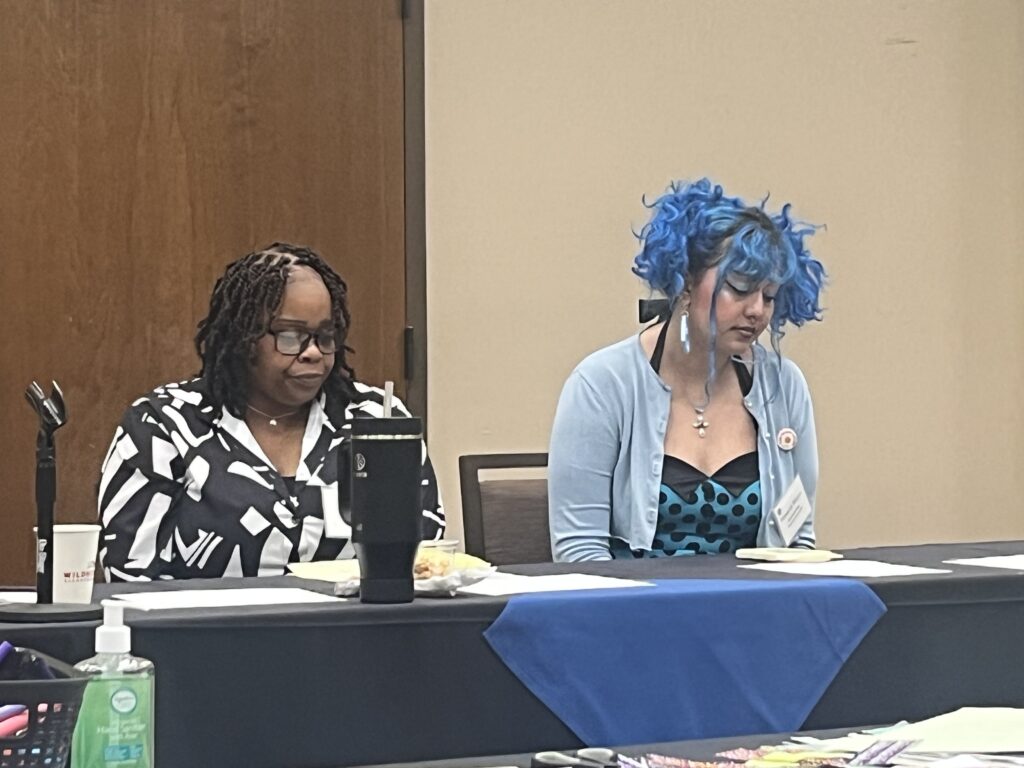 Two people sit at a conference table during a community discussion or workshop, looking down at notes and materials in front of them.