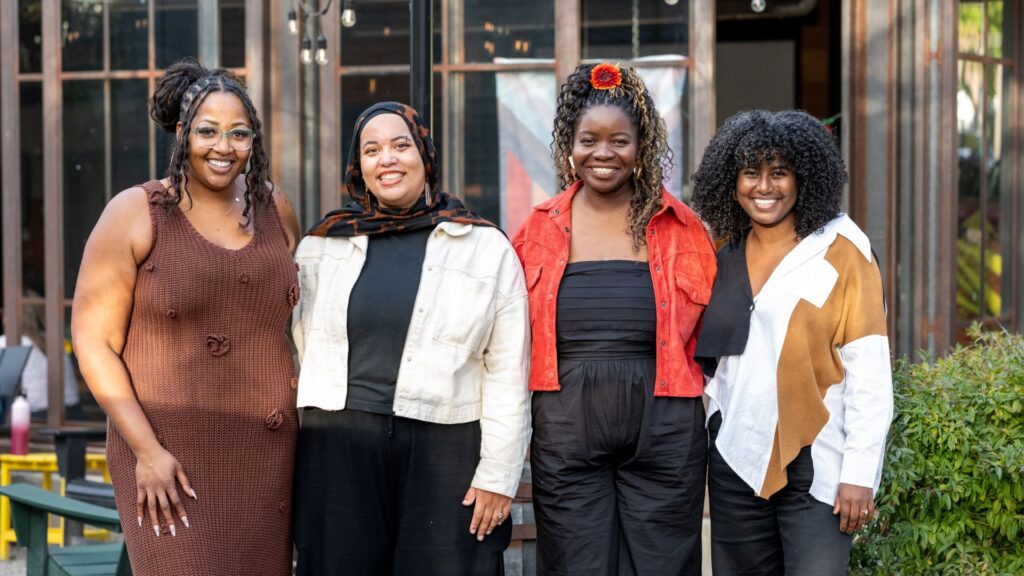 Four members of Social Justice Fund Northwest's Directors Team stand side by side outdoors, smiling at the camera in front of a building with large windows and string lights.