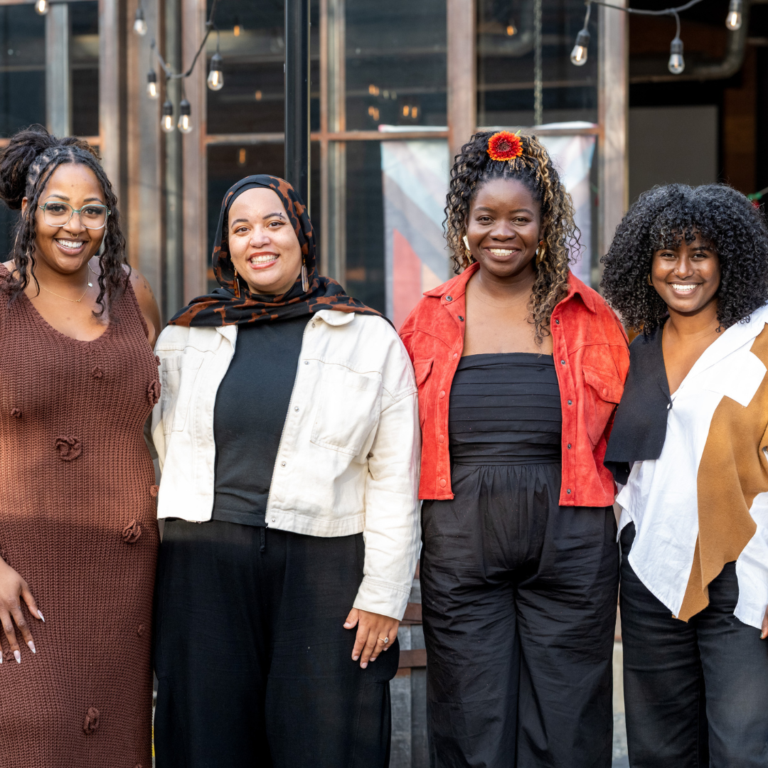 Four members of Social Justice Fund Northwest's Directors Team stand side by side outdoors, smiling at the camera in front of a building with large windows and string lights.